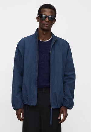 Uomo con occhiali da sole scuri, giacca leggera blu scuro con zip sopra un maglione scuro e pantaloni neri su uno sfondo semplice.