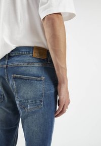 Jeans de mezclilla azul con un parche de cuero en la parte posterior de la cintura, cinco bolsillos y una textura ligeramente desgastada. Camiseta blanca visible.