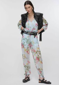 Femme portant un chemisier transparent à fleurs et un pantalon, ceinture noire ornée de clous, gilet en cuir noir, bracelets argentés, et sandales noires à clous, debout les mains sur les hanches.