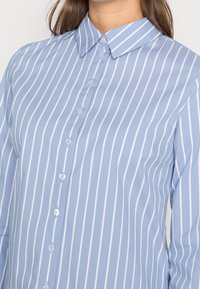 Camisa de botón azul claro con rayas verticales blancas, cuello clásico y botones visibles. Hecha de una tela suave con un corte entallado.