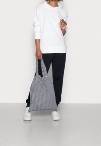 Biała bluza z kapturem i sznurkami, zestawiona z granatowymi dresami. Osoba trzyma torbę na zakupy w paski w czarno-białe pionowe paski.