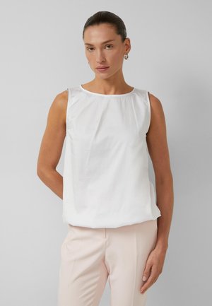 Frau mit zurückgebundenem dunklem Haar, die eine ärmellose weiße Bluse und hellbeige Hose trägt und vor einem einfarbigen hellgrauen Hintergrund steht.