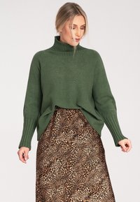 Zielony dzianinowy sweter z wysokim kołnierzem i prążkowanymi mankietami, w połączeniu z brązową spódnicą w panterkę o zwiewnym kroju.