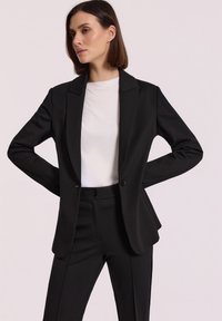 Kobieta w czarnym, dopasowanym żakiecie i pasujących spodniach, z białą bluzką, stojąca z rękami na biodrach na jednolitym tle.