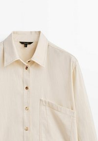 Camicia color crema con colletto a punta, taschino frontale e patta con cinque bottoni tondi. Tessuto liscio e leggero.
