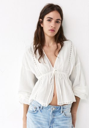 Jonge vrouw met lang bruin haar die een losse witte blouse draagt, geknoopt in de taille, en lichtblauwe high-waisted jeans tegen een effen achtergrond.