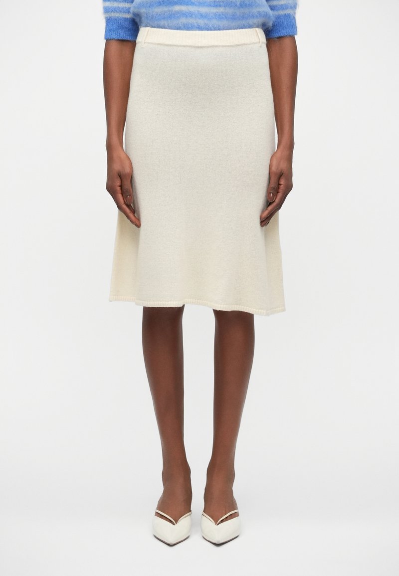 Jupe évasée en tricot couleur crème, présentant une texture douce, une taille ajustée et des détails de couture subtils. Associée à des escarpins blancs à bout pointu.