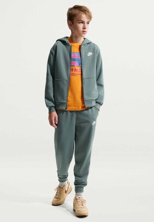 Jongen staat, draagt een groen Nike trainingspak, oranje T-shirt met print, beige sneakers, kijkt naar de zijkant met handen in de zakken.