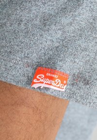 Lähikuva harmaasta kankaasta, jossa on teksturoitu pinta. Laidassa on oranssi "SuperDry" -tunnus ommeltuna, ja se tuo brändin nimen selkeästi esiin.