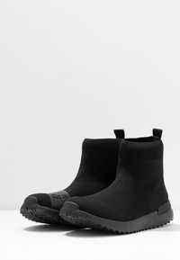 Black knitted ankle boots with a sock-like upper, rubber sole, and textured tread. Features pull tabs for easy wear.