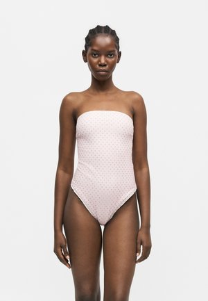 Tânără femeie de culoare cu părul împletit, purtând un costum de baie roz deschis fără bretele cu buline roșii mici, stând pe un fundal simplu.