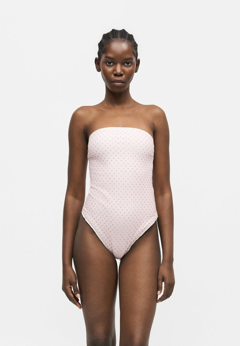 Νεαρή μαύρη γυναίκα με πλεξούδες, που φοράει ένα ροζ ολόσωμο μαγιό χωρίς τιράντες με μικρές κόκκινες πουά, στέκεται μπροστά σε ένα απλό φόντο.