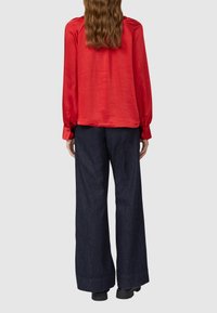 Frau mit langem, welligem Haar, die eine rote Langarmbluse und dunkle, weit geschnittene Hosen trägt, von hinten vor einem grauen Hintergrund betrachtet.