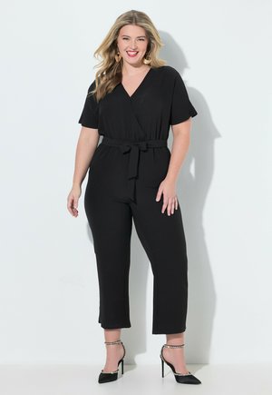 Hymyilevä nainen, jolla on vaaleat hiukset, musta vyötäröllä varustettu haalari ja mustat nilkkahihna korkokengät, seisomassa valkoista taustaa vasten.