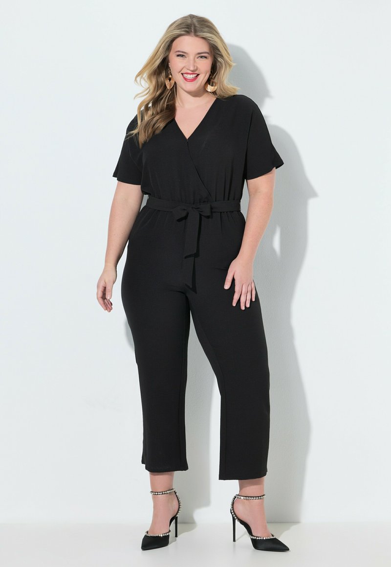 Mujer sonriente con cabello rubio, vestida con mono negro con cinturón y tacones altos negros con tiras en el tobillo, de pie frente a un fondo blanco.