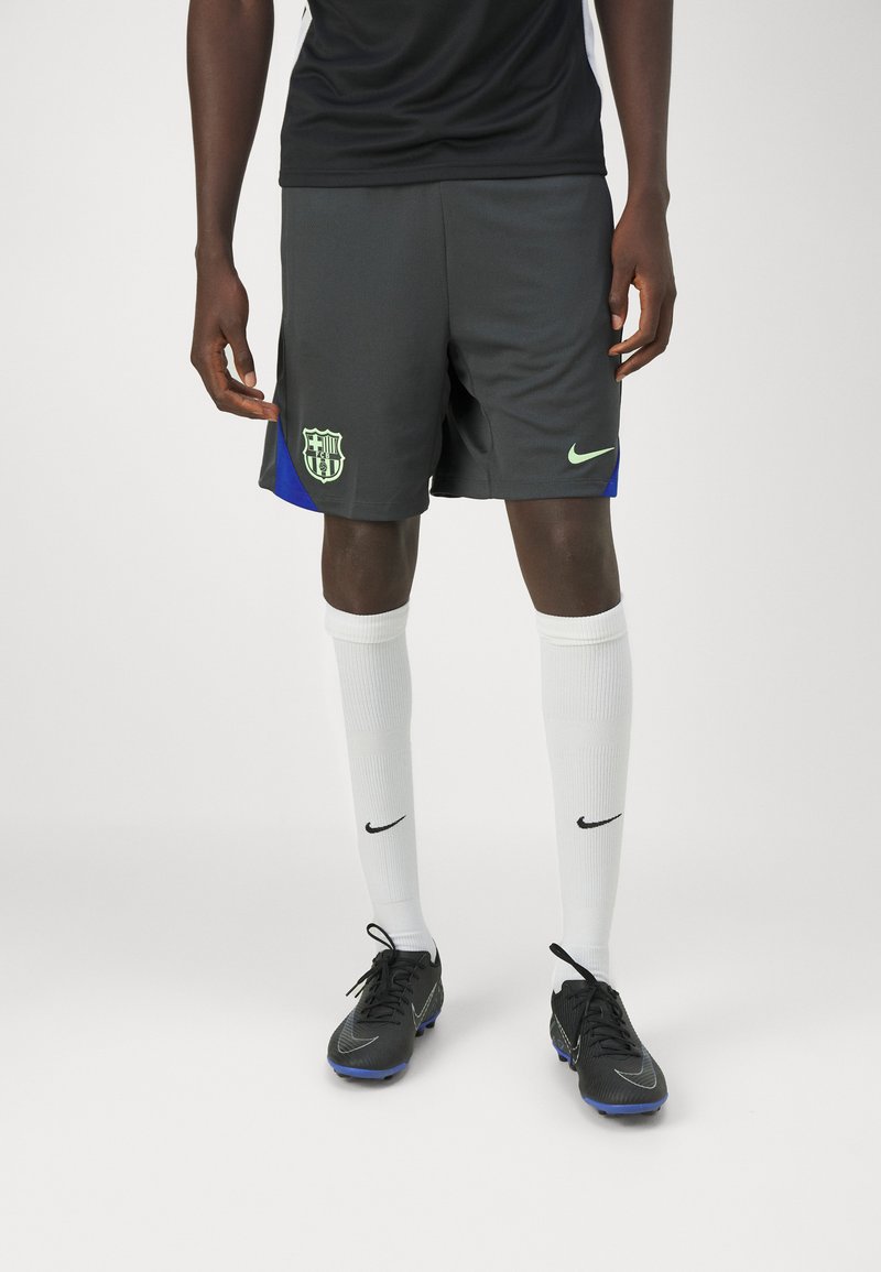 Pantalones cortos de fútbol grises con un logo de Barcelona en neón verde y acentos laterales azules, combinados con calcetas blancas y botas Nike negras.