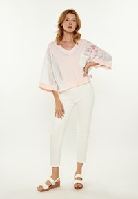 Lichtroze blouse met bloemaccenten en opengehaakte mouwen, gecombineerd met witte aangesloten broek en witte sleehakken.