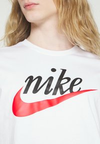 T-shirt bianco con logo "nike" nero e grafica swoosh rossa, colletto rotondo, maniche corte e texture in cotone morbido.