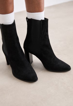 Bottines en daim noir avec un bout pointu, un talon carré et des panneaux latéraux élastiques. Le design présente une esthétique minimaliste et une texture lisse.