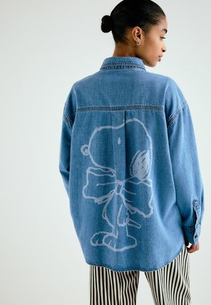 Frau trägt ein locker sitzendes blaues Jeanshemd mit einer großen weißen Umrisszeichnung eines Cartoon-Charakters auf dem Rücken, kombiniert mit schwarz-weiß gestreiften Hosen.