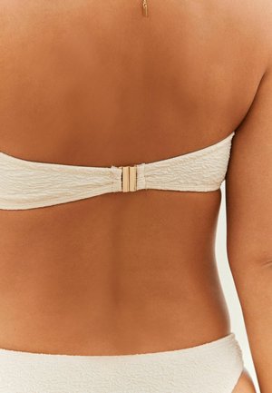 Cremefarbener Bikini-Oberteil aus strukturiertem Stoff, mit einem goldenen Verschluss hinten. Das Design ist trägerlos und flach im Rücken.