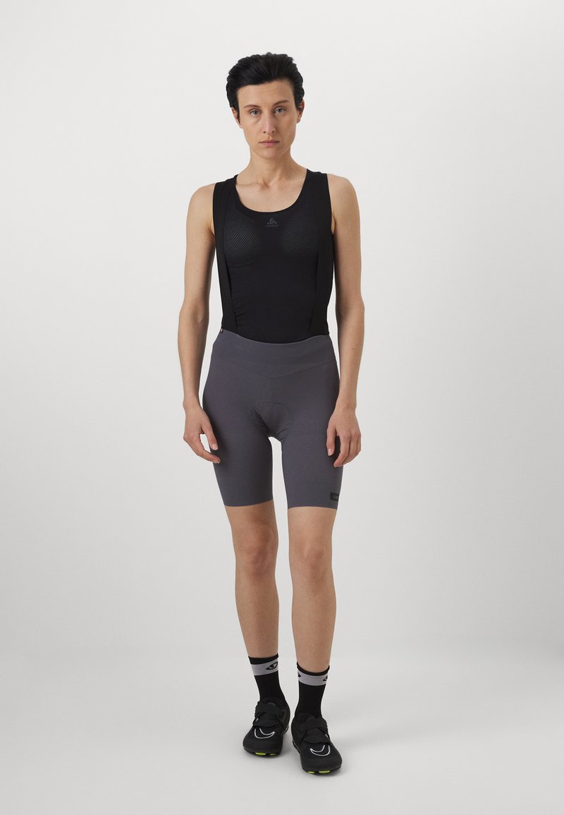 Salopette nera da ciclismo con un design aderente, caratterizzata da pannelli in rete e pantaloni corti imbottiti grigi. Scarpe da ciclismo nere e calzini a righe completano il look.