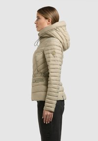 Vrouw in beige gewatteerde donsjas met capuchon, staand in profiel tegen een effen achtergrond, met zwarte broek.