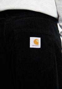 Zwarte corduroy broek met achterzak en witte Carhartt logo patch, gedragen met een lichtgrijze top die zichtbaar is bij de taille.