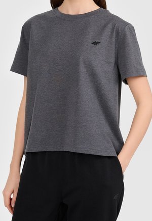 Personne portant un t-shirt gris foncé à manches courtes avec un petit logo noir "4F" sur la poitrine et un pantalon noir, main dans la poche.