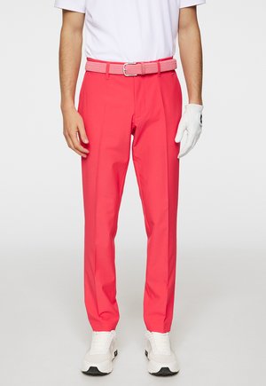Ljusa rosa golfbyxor med en slät textur, utrustade med ett vävt bälte och klassisk rak passform. Bär vit golfhandskar och sneakers.