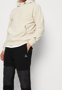 Cremfarbener Fleece-Hoodie mit Fronttasche und schwarzem Jordan-Logo auf dem Ärmel. Kombiniert mit schwarzen strukturierten Hosen mit Reißverschlusstasche und Logo.