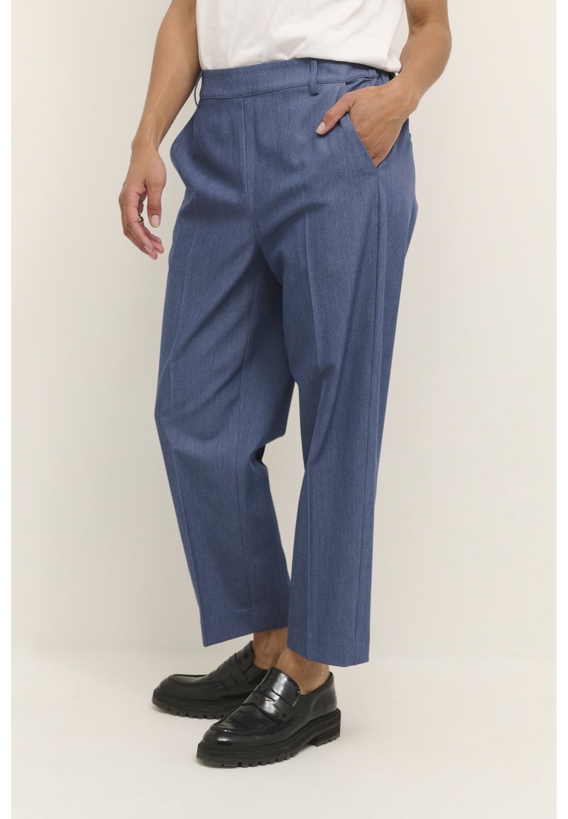 Personne portant un pantalon bleu sur mesure court, une chemise blanche et des mocassins noirs, avec une main dans la poche devant un fond uni.