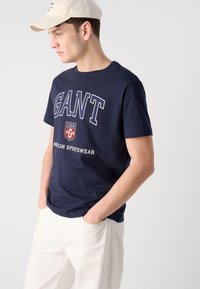 Tricou din bumbac bleu marin cu logo-ul "GANT" în alb, design cu guler rotund și mâneci scurte. Asortat cu pantaloni albi și o șapcă bej.