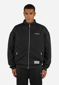 Sort lynlås sweatshirt med høj krave, udstyret med sidelommer og broderet logo på brystet; lavet af blødt, tekstureret stof.