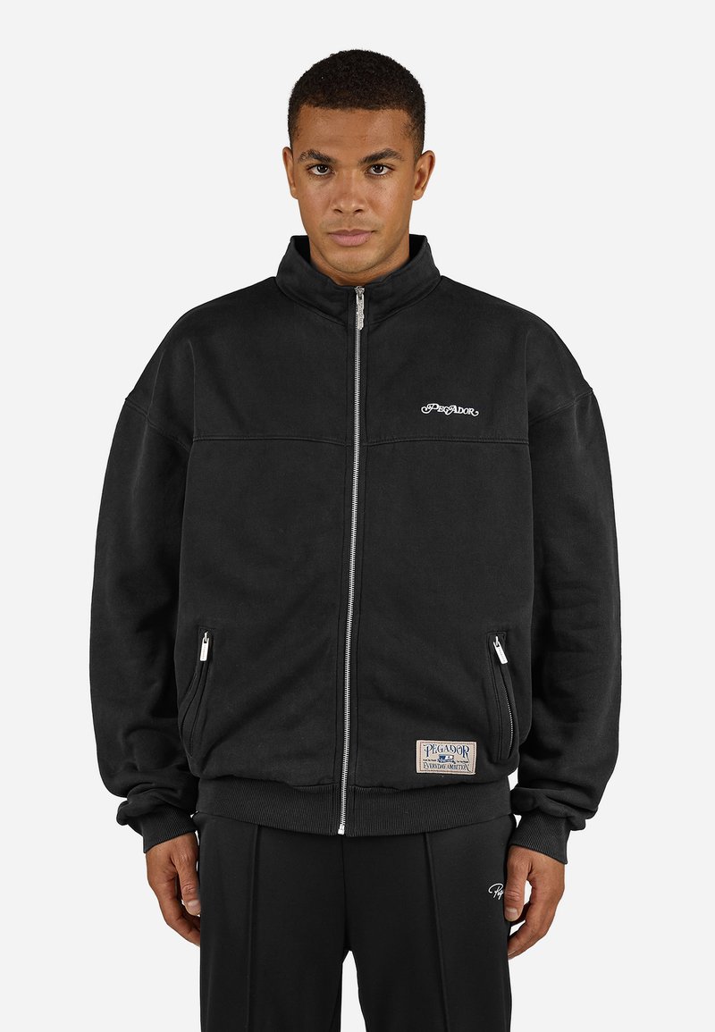 Veste à zip noire avec un col haut, dotée de poches latérales et d'un logo brodé sur la poitrine ; fabriquée en tissu doux et texturé.
