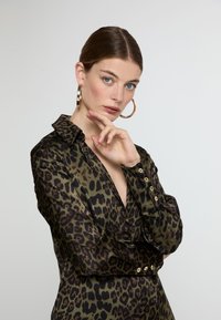 Leopard print blouse in olive green with a deep V-neck, buttoned cuffs, and glossy finish. Model poses with a hand on chin, gold hoop earrings.