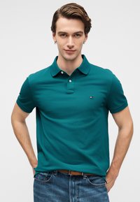 Πράσινη μπλούζα πόλο από βαμβακερό μείγμα, με γιακά, τρία κουμπιά και μικρό λογότυπο στο αριστερό στήθος. Φοριέται με μπλε τζιν.