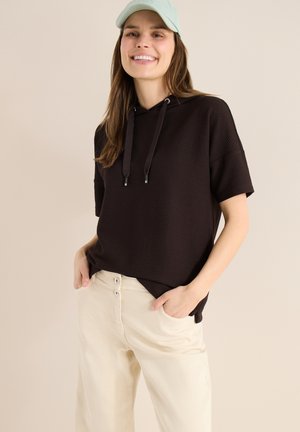 Ung kvinde iført en sort kortærmet hættetrøje, beige bukser og en lyseblå baseballkasket, smilende med hænderne i lommerne.