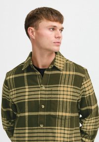 Grøn og beige ternet flannelskjorte med spids krave, knaplukning foran og brystlomme. Tekstureretstof med markante vandrette og lodrette striber.