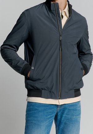 Person iført en mørkegrå lynlåstrøje med lommer og ribbede manchetter, kombineret med lyseblå jeans og en beige skjorte under.