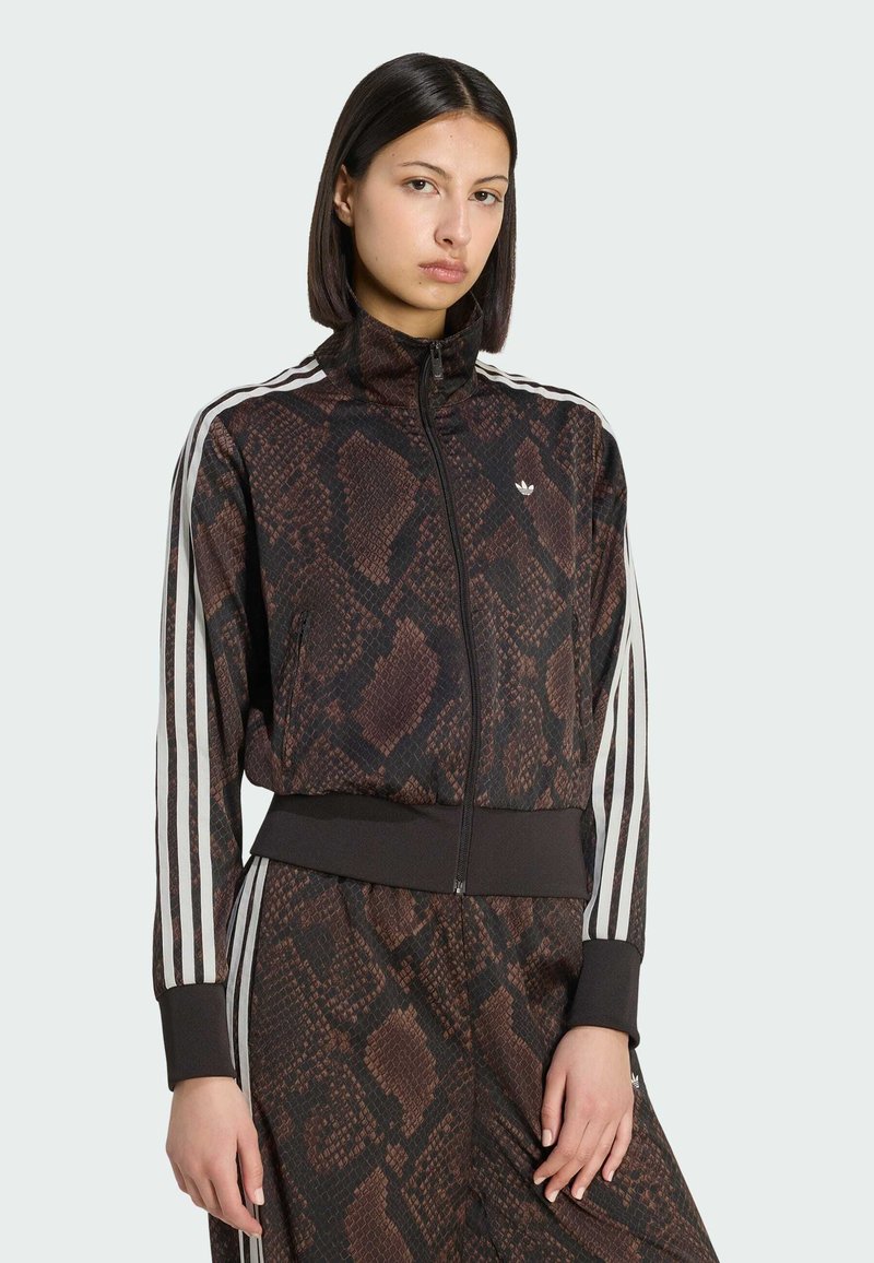 Jeune femme portant une veste marron à motif serpent avec fermeture éclair et un pantalon assorti avec des bandes blanches le long des manches et des jambes, debout devant un fond blanc.