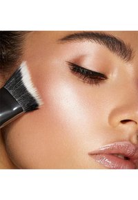 Meikkisivellin, jossa on musta varsi ja pehmeä, valkoinen harjaspää, levittää meikkiä hohtavalle, sileälle iholle, jossa on luonnollisia korostuksia.