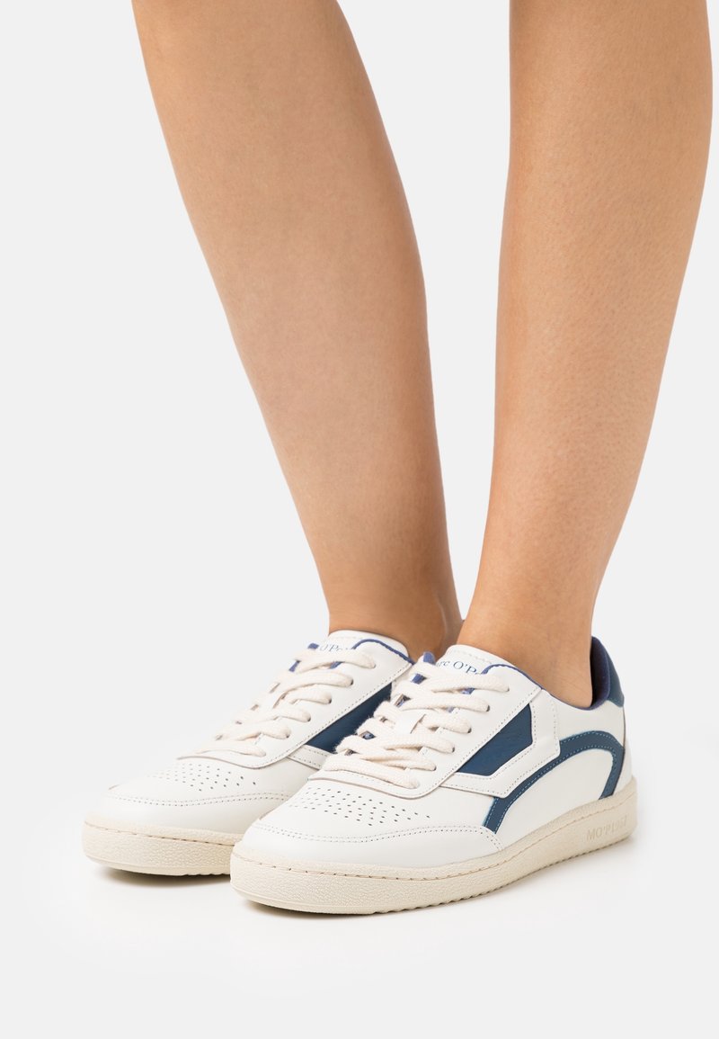 Baskets blanches avec accents bleu marine portées pieds nus, présentant un design perforé à l'avant et une fermeture à lacets.