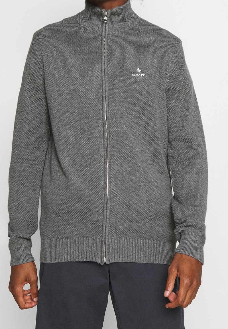 Sudadera gris de punto con cremallera y cuello alto, puños y dobladillo acanalados, que presenta un pequeño logo blanco en el lado izquierdo del pecho.
