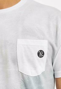 Valge puuvillane T-särk, millel on rinnatasku, millel on ümmargune must logo, delikaatne tekstuur ja ümar kaelus.