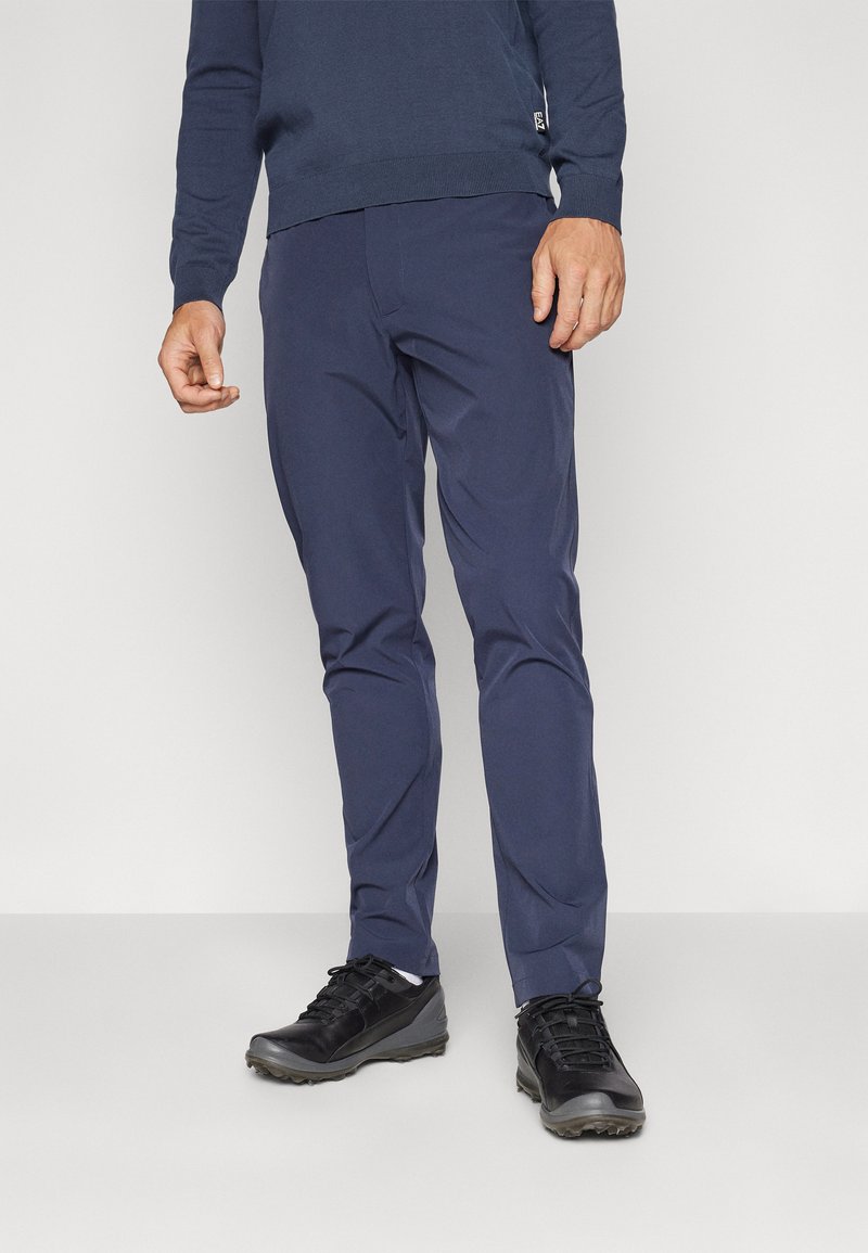 Pantalons bleu marine au design ajusté, fabriqués dans un tissu lisse. Associés à des chaussures noires avec une semelle texturée et des lacets.