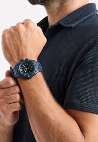 Montre bleue avec un bracelet texturé, cadran noir contrastant, marqueurs blancs et accents orange. Portée sur un avant-bras bronzé.