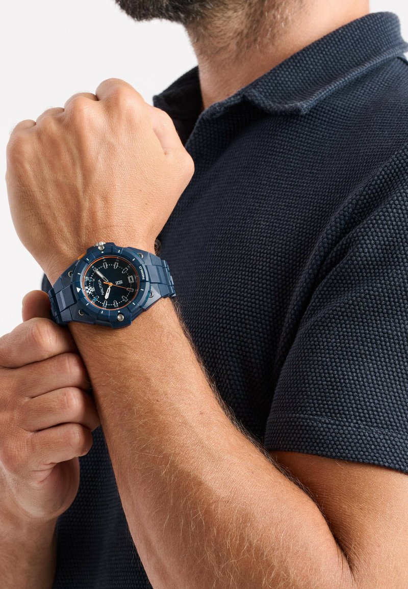 Montre bleue avec un bracelet texturé, cadran noir contrastant, marqueurs blancs et accents orange. Portée sur un avant-bras bronzé.
