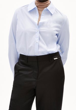 Osoba ubrana w jasnoniebieską zapinaną na guziki koszulę na beżowym golfie, czarne, wysokie, dopasowane spodnie, stojąca na białym tle.