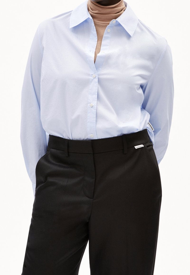 Personne portant une chemise bleu clair boutonnée sur un col roulé beige avec un pantalon noir sur mesure taille haute, debout devant un fond blanc.